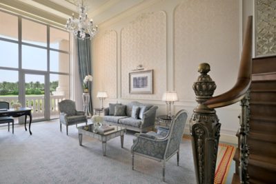 This image showcases the opulent interior of a hotel suite, likely part of the Accor brand. The room is furnished with a light blue and white patterned sofa, accompanied by two matching armchairs. A mirrored coffee table sits in the center, holding a book and a vase of white flowers.  Two end tables with lamps flank the sofa. A writing desk with a chair is positioned near the large windows, which offer a view of a landscaped area with palm trees, suggesting a warm climate location.  The room's walls are adorned with elegant wallpaper featuring a subtle damask pattern. A framed picture hangs on the wall, depicting a classical building, adding to the sophisticated ambiance. An elaborate crystal chandelier hangs from the high ceiling, and a grand staircase with a wooden handrail and ornate metal balusters is partially visible. The flooring is covered with a patterned carpet in soft blue and white hues. The overall atmosphere suggests a luxurious leisure context.