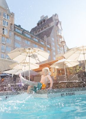 boy sitting on the side of the pool splashing water infront of lounge chairs with umbrellas with a large building in the background