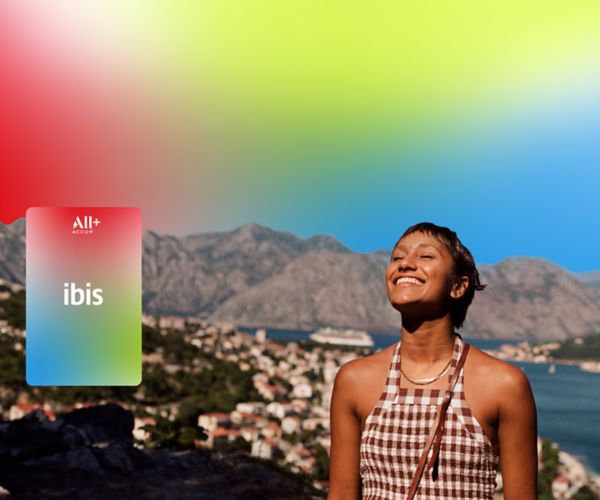A smiling woman , closing eyes, in front of a mountain landscape. The background is a red and green gradient, the ALL Accor+ ibis subscription colors that grants -15% off your in hotel stays all year long !