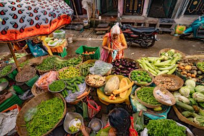 A bustling street scene unfolds from an overhead perspective, showcasing a vibrant array of fresh produce displayed at a market stall. Two women tend to their wares, their faces partially obscured, amidst overflowing baskets of vibrant green vegetables, plump orange pumpkins, and ripe yellow melons. The woman on the left, adorned in a colorful sari, expertly weighs a customer's selection of green limes, her hands moving with practiced ease. Meanwhile, another woman, her back to us, stands amidst the colorful chaos, organizing the various fruits and vegetables.