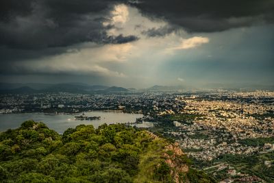 A sprawling cityscape stretches out below a dramatic, cloud-filled sky.  Lush green foliage covers the hillside in the foreground, offering a vantage point for taking in the urban expanse and a glimpse of the water beyond.  The cityscape is a mix of white and beige buildings, punctuated by the occasional high-rise.  The water is a deep blue, reflecting the overcast sky.