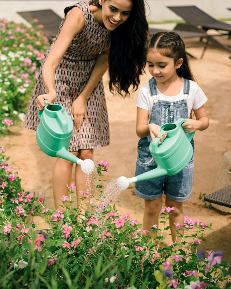 A smiling woman and a young girl watering pink flowers in a garden with green watering cans.