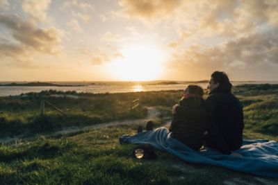 Un couple assis sur un plaid, contemple le coucher du soleil lors d’un pique-nique en amoureux en Bretagne