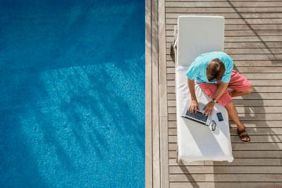 Un homme travaille sur son ordinateur portable, assis sur un transat au bord de la piscine d’un hôtel