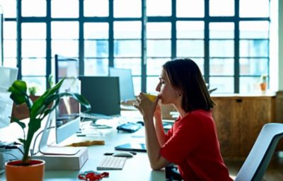 Une femme portant un top rouge boit une boisson chaude pour s’accorder un temps de repos au travail, devant son ordinateur dans l’open space
