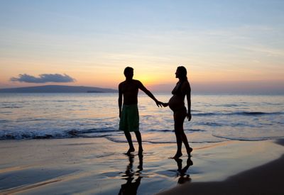 An expectant couple silhouetted against a beach sunset, enjoying a babymoon