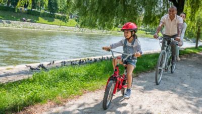 A smiling child cycling with parents along a tree-lined waterfront in a sunny park