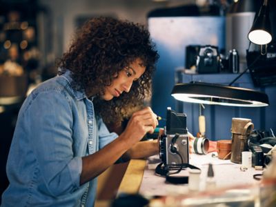 Une femme entrepreneure qui répare un ancien appareil photo à l’aide d’un mini-tournevis, assise à son bureau dans son atelier 