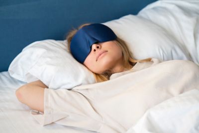 A person wearing a blue satin sleep mask lying comfortably in bed
