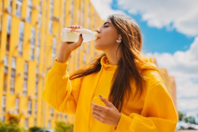 Une jeune femme portant un sweat jaune boit de l’eau dans la rue, devant un bâtiment à la façade jaune