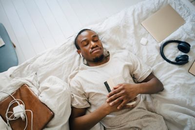 Mann liegt auf dem Rücken im Bett mit einem Buch auf seinem Bauch und Laptop neben sich