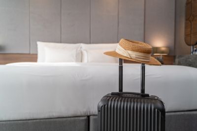 A suitcase and a straw hat in front of a comfortable bed in a hotel room