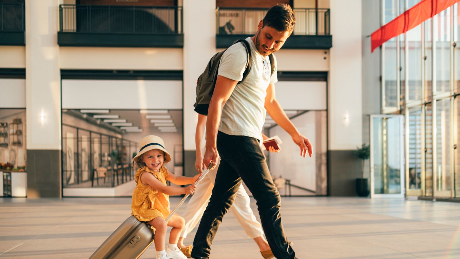 Padre llevando a su hija pequeña sobre una maleta de ruedas en lo que parece un aeropuerto