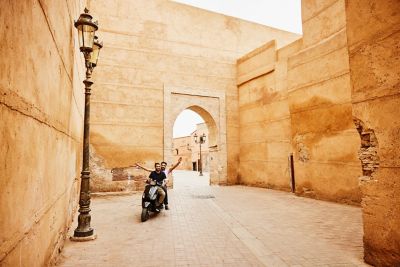 Two smiling friends on a motorbike amid the sandstone walls of Marrakech Medina