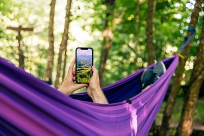 A person lying in a purple hammock in the woods holding their phone up