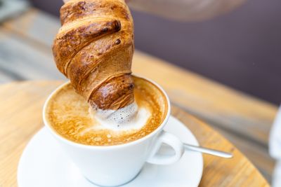 A croissant being dunked into a cup of froth-topped coffee