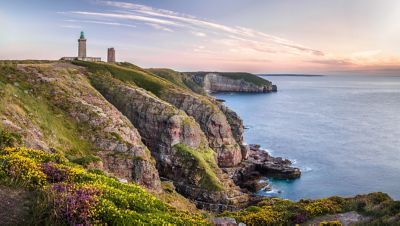 Phare du cap Fréhel au crépuscule, sur une falaise verdoyante parsemée de fleurs sous un ciel bleu aux teintes rosées