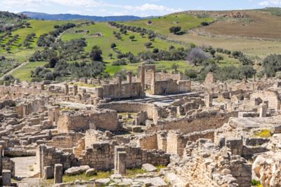 Ruinas romanas antiguas de piedra en Dougga, Túnez, con colinas verdes y cielo azul de fondo