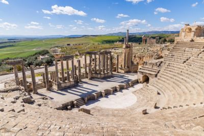 Ruinas del teatro romano de Dougga en Túnez, con columnas y gradas de piedra bajo un cielo azul con nubes
