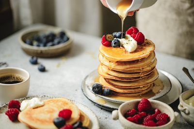 Maple syrup being poured over a stack of fluffy, berry-topped pancakes