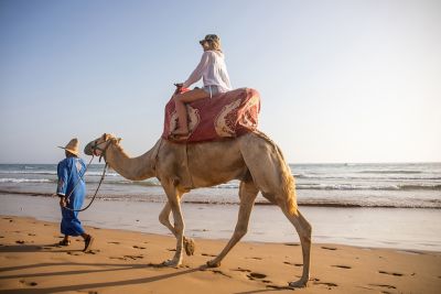 A woman riding on a camel led by its owner on a calm, sandy beach in Morocco