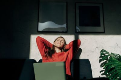 Une femme portant un top rouge s’étire au soleil pour faire une pause pendant sa journée de télétravail, installée devant son ordinateur portable à l’extérieur