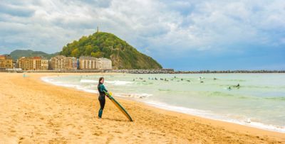 Surfista en playa de Zurriola, con edificios y monte de fondo, con el cielo nublado