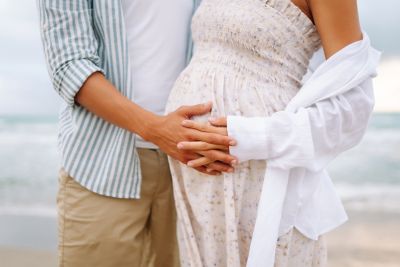 Expectant parents on a babymoon, dressed in light colours and standing on a beach