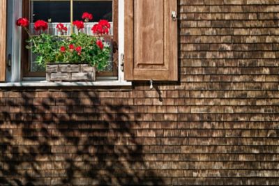 Fassade eines historischen Bauernhauses mit dunklen Holzschindeln und roten Blumen vor dem Fenster
