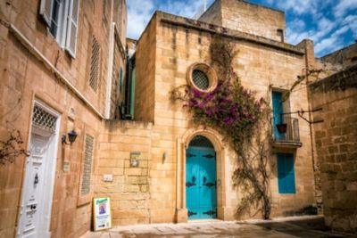 Una calle estrecha con edificios de piedra, una puerta turquesa y flores moradas en Mdina, Malta