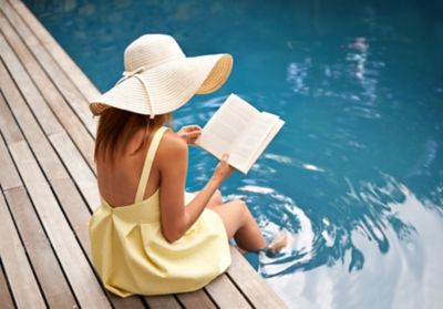 Une femme portant une robe jaune et un chapeau, assise au bord d’une piscine et qui se détend en lisant un livre les pieds dans l’eau.