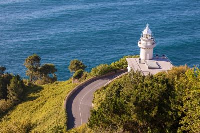 Faro blanco sobre acantilado verde junto al mar azul, con carretera sinuosa