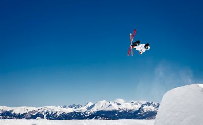 Esquiador en un salto acrobático contra un cielo azul claro y montañas nevadas