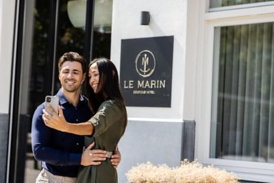 Un couple heureux qui prend un selfie devant la devanture de l’hôtel Le Marin Hotel Rotterdam City