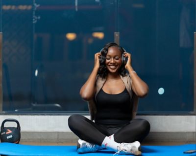 Une femme est assise en tailleur sur un tapis de fitness bleu, ajustant son casque audio pour se concentrer en écoutant de la musique