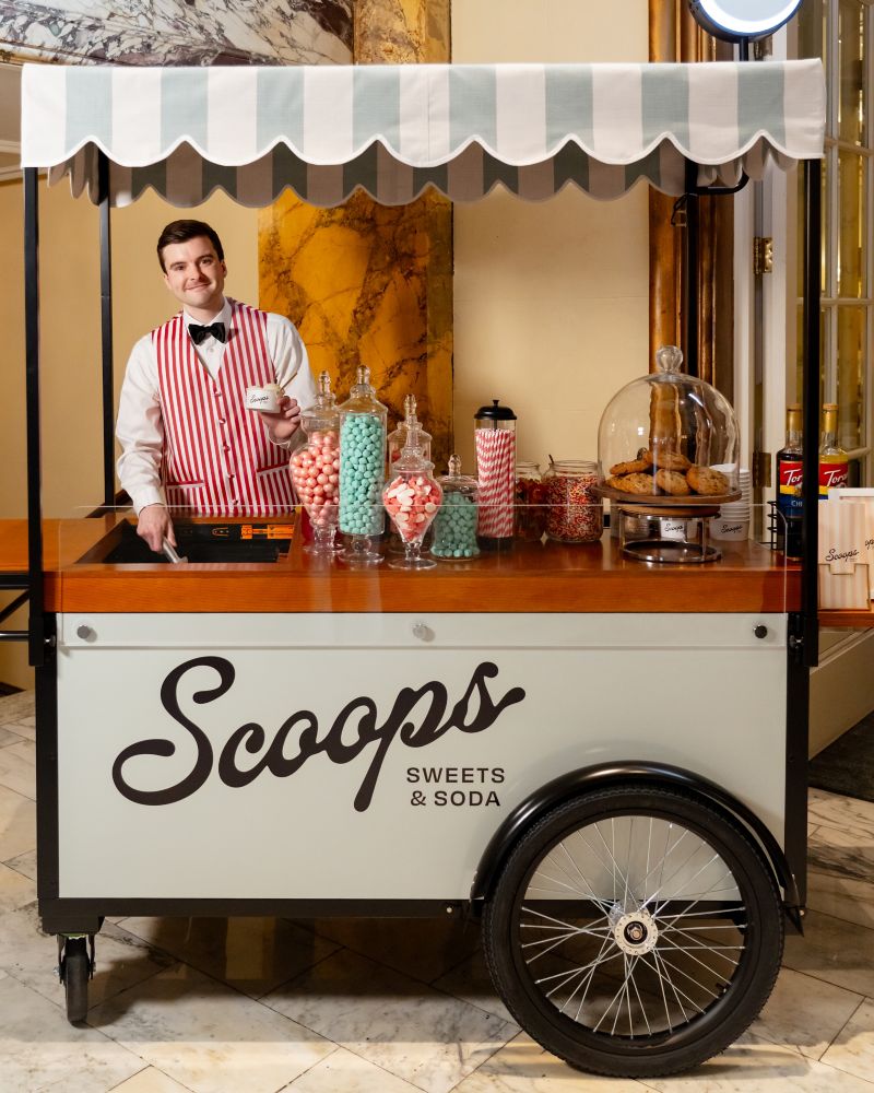 Man offering ice cream from an ice cream cart that says scoops sweets & soda