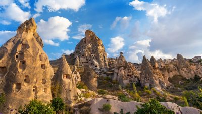 This captivating image showcases the unique landscape of Cappadocia, Turkey, with its distinctive rock formations.  The image features a series of cone-shaped rock structures, many with carved openings suggesting ancient dwellings or storage spaces. Some of these structures are quite large, dominating the foreground, while others cluster together in the background. The rocks vary in height and shape, creating a visually interesting composition. The terrain surrounding the rocks is dry and appears sparsely vegetated with some shrubs and small trees.  A vibrant blue sky dotted with white and light grey clouds provides a striking backdrop to the earthy tones of the rock formations. There are no visible people in the image.  The sunlight casts a warm glow, enhancing the colors and textures of the scene. There are no discernible logos, brands, or text. No Accor branding elements are visible, nor any evidence of partnerships like PSG.  There are no shops depicted in the scene.  The context is unclear; it could be either business or leisure. No specific event is apparent, and the time of day appears to be late afternoon. The overall impression is one of natural beauty and historical significance.