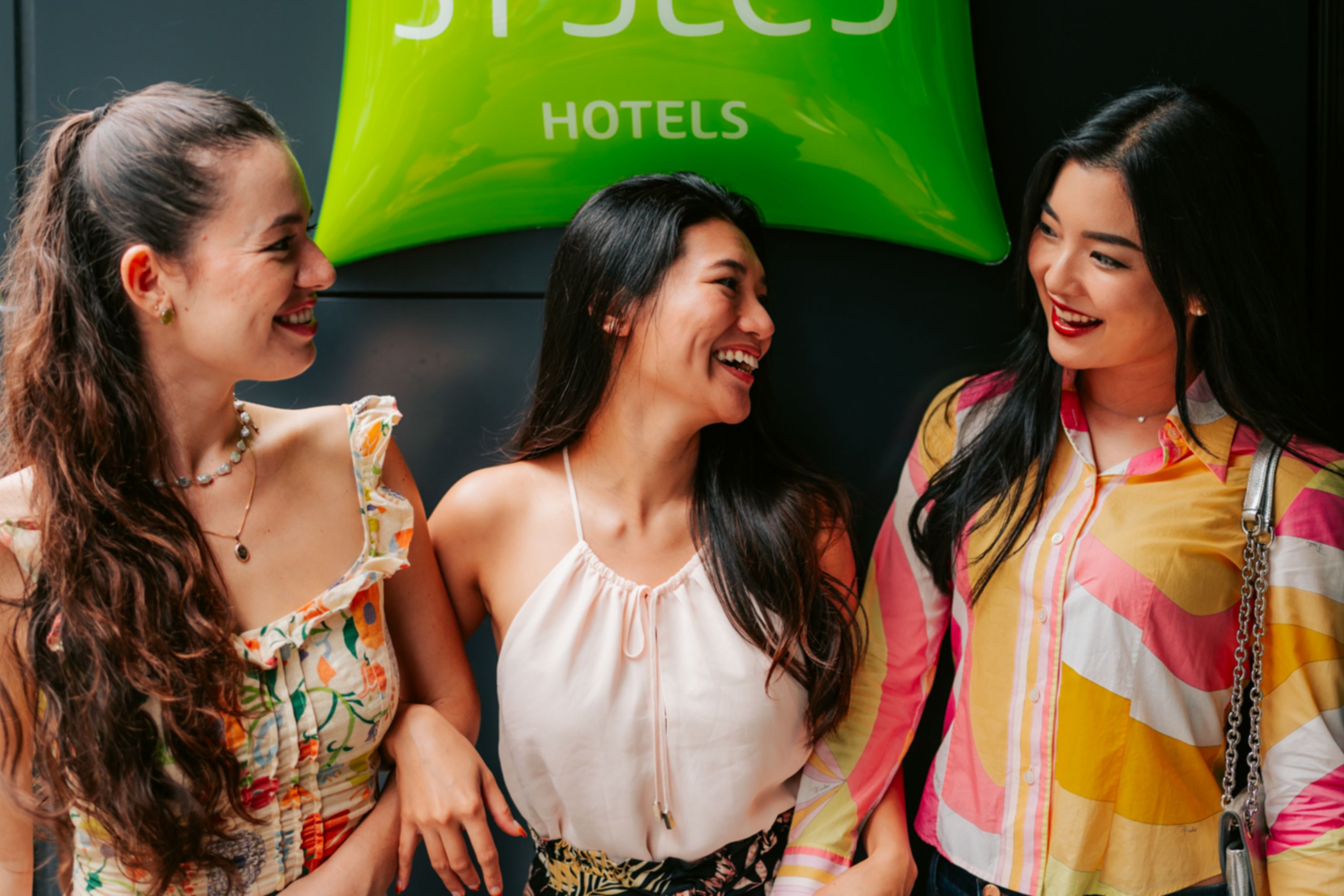 Three women laughing together in front of an ibis Styles sign outside a hotel in Nagoya, Japan