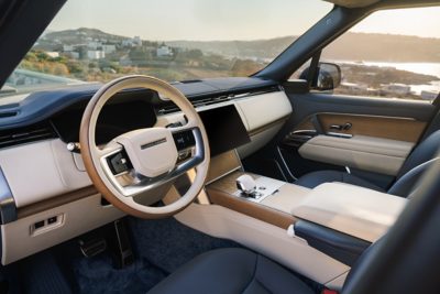 The interior of a luxurious Range Rover vehicle is shown, featuring cream-colored leather seats and a matching dashboard.  The wood grain accents on the steering wheel and door panels add a touch of sophistication. The car is parked facing a picturesque coastal town bathed in the warm glow of the setting sun. The sky is clear with a few wisps of clouds, and the tranquil sea stretches out in the distance.