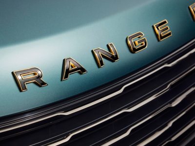 Close-up of the Range Rover emblem, crafted in bold, gold lettering against a sleek, teal car hood. The smooth, reflective surface of the hood highlights the luxurious craftsmanship of the vehicle. Below the emblem, the car's grill features a striking contrast of black and silver, further enhancing the vehicle's modern and sophisticated aesthetic.