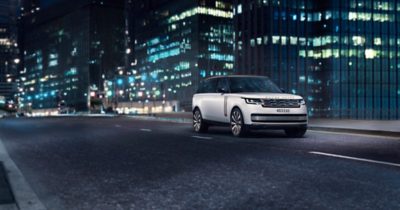 A white Range Rover SUV drives down a city street at night, illuminated by the glow of streetlights and surrounding buildings.  The Range Rover, with its sleek design and copper accents, stands out against the backdrop of a bustling city. 