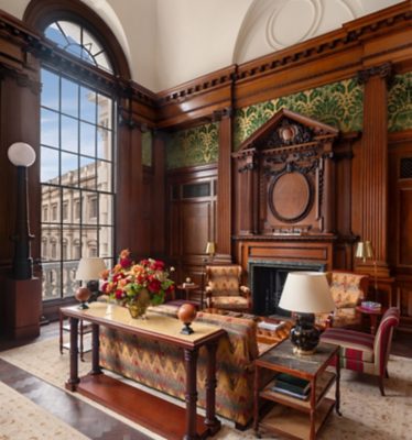 This image showcases a luxuriously appointed sitting area within a building, possibly a high-end hotel or private residence affiliated with Accor. The room is richly decorated with dark, polished wood paneling and ornate architectural details, including a large arched window that offers a glimpse of a neighboring building's facade and a partially visible sky.  A large floral arrangement in a glass vase sits on a long, narrow table with a patterned fabric skirt and turned wooden legs, positioned in front of a sofa upholstered in a similar patterned fabric.  Two armchairs, one with a chevron pattern and the other with vertical stripes in red, gold, and cream, flank the sofa. A small, round wooden ottoman rests on the patterned rug in the center of the seating area.  A side table with a dark, possibly ceramic lamp and a book adds to the sophisticated atmosphere. The fireplace, framed by the intricate woodwork, has a dark green marble surround. The walls feature sections of green and gold damask wallpaper, further enhancing the opulent aesthetic.  The high ceiling and large window contribute to a sense of spaciousness and grandeur. The overall mood suggests a setting designed for leisure, though it could also serve as a business meeting area. No people are visible in the scene.  The room is well-lit, likely with a combination of natural light from the window and light from the lamps.
