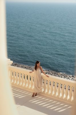 A woman, wearing a flowing, light-colored, ankle-length dress and sandals, leans against the balustrade of a balcony, overlooking a tranquil expanse of blue ocean. Her expression is serene as she gazes towards the right side of the frame.  The balcony features a classic, cream-colored balustrade with repeating balusters, casting long shadows across the tiled floor.  The rocky shoreline is just visible beyond the balcony. The sky is clear and the light suggests a daytime scene.  The overall impression is one of relaxed leisure, possibly at an upscale seaside resort.
