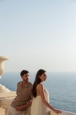 A man and a woman stand on a balcony overlooking a vast, tranquil expanse of ocean. The clear, pale blue sky provides a serene backdrop for the scene. The man, wearing a light brown, textured, short-sleeved polo shirt and light-colored pants, leans casually against the balcony railing. He looks directly at the camera with a neutral expression. Beside him, the woman, dressed in a flowing, light cream-colored sundress, gazes out at the sea, a contemplative expression on her face. The balcony features a decorative urn, adding a touch of elegance to the setting.  The overall atmosphere suggests a leisurely vacation moment. No visible logos or branding are present.
