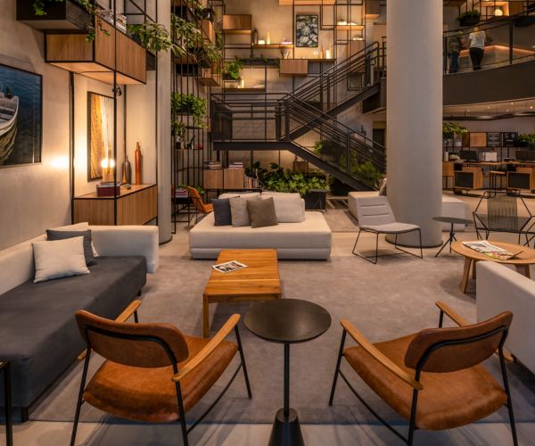 A modern open-plan office space with a lounge area that includes sofas with cushions, wooden coffee tables, and contemporary chairs. A staircase leads to an upper level, adorned with career information displays. Potted plants add a touch of greenery while office desks and computers are visible in the background.