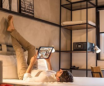 A person lies on a bed with high-quality bedding, their legs resting against the wall, holding a tablet and looking at it. The room, reminiscent of ibis hotels, has minimalistic decor with shelves, a safe, a window, and a desk with a laptop and lamp in the background—a perfect setting for restful sleep.