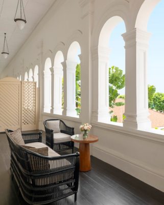 A serene hallway adorned with a sequence of graceful arches, leading to a vista of lush greenery. The space is tastefully furnished with a black wicker sofa and armchair, complemented by a round wooden table. Atop the table sits a vase filled with a delicate arrangement of pink and white flowers.  The bright interior is illuminated by natural light streaming through the arched windows, highlighting the white walls and dark wooden floor. The overall ambiance exudes tranquility and elegance, suggesting a luxurious and comfortable experience. The scene appears to be set in a context of leisure, offering a peaceful retreat amidst a tropical setting.