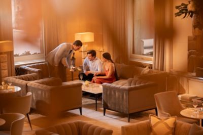 A couple is served champagne by a hotel staff member in an elegant, warmly lit lounge.