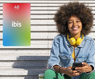 A smiling person with a large afro hairstyle, wearing a denim jacket and yellow headphones around their neck, sits on steps. To the left, there is an Ibis logo on a gradient background with red and green hues. Perfect for leisure or business stays, enjoy 15% OFF your next visit!