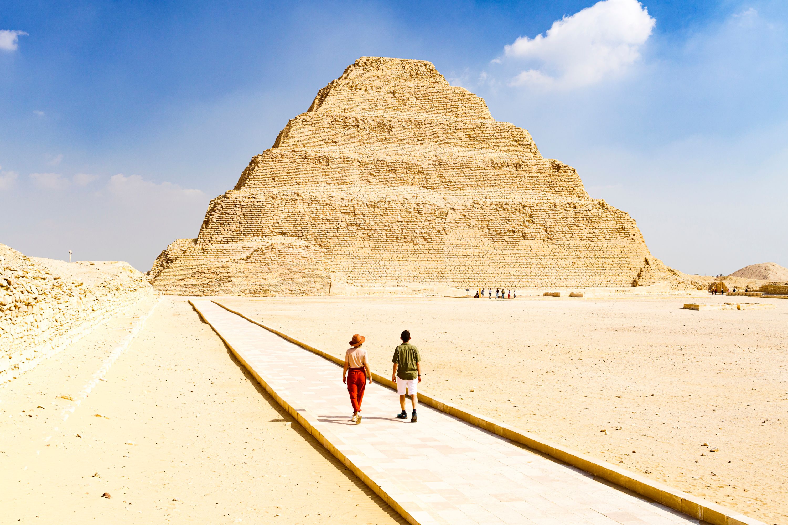Man and woman walking on a pedestrian path towards the Step Pyramid of Djoser under a bright clear sky, Saqqara, Egypt.